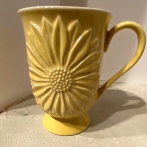 May include: A yellow ceramic mug with a raised sunflower design. The mug has a handle and a pedestal base.
