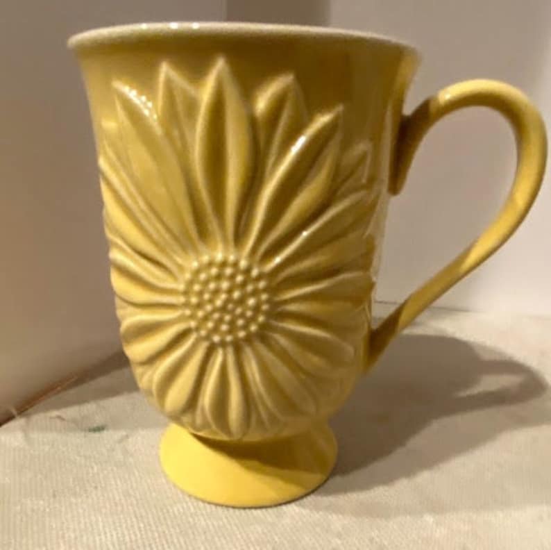 May include: A yellow ceramic mug with a raised sunflower design. The mug has a handle and a pedestal base.