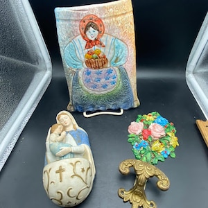 May include: Three ceramic wall plaques featuring religious imagery. The first plaque depicts a woman in a red and white dress holding a basket of fruit. The second plaque depicts a woman in a blue dress holding a child. The third plaque depicts a floral design with pink, yellow, and blue flowers.