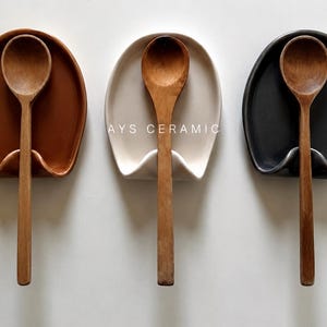 Può includere: Tre poggiacucchiaio in ceramica marrone, crema e nero, ciascuno con un cucchiaio di legno. I poggiacucchiaio hanno un design curvo con uno slot per il manico del cucchiaio. Il testo "AYS CERAMIC" è visibile sul poggiacucchiaio color crema.