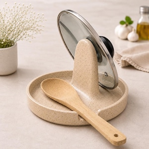 Puede incluir: Un reposautensilios de cocina beige moteado con un soporte central vertical que sostiene una tapa de olla circular de vidrio con borde de metal y pomo negro. Una cuchara de madera clara descansa horizontalmente sobre la base de la bandeja.