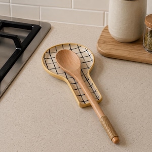 May include: A wooden spoon rests on a yellow spoon rest with a black grid pattern. The spoon rest is on a light-coloured countertop next to a hob, a wooden chopping board, and jars.