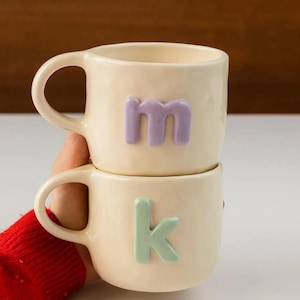 Può includere: Due tazze in ceramica color crema impilate. La tazza superiore ha una "m" lavanda e quella inferiore una "k" verde menta. Ogni tazza ha un manico curvo. Ideali per il caffè.