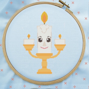 May include: A cross-stitch embroidery hoop with a light blue background featuring a yellow candle holder with a smiling face. The candle holder has three candles, one large and two smaller candles.