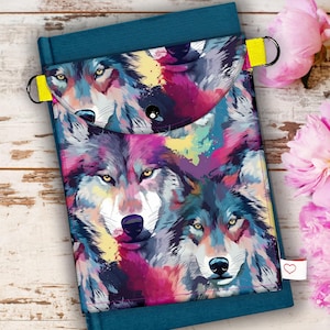 Peut inclure: Une couverture de livre turquoise avec un rabat orné d'un motif de loup coloré. L'œuvre d'art comprend des nuances de violet, de bleu et de jaune. La couverture a des boucles jaunes et une petite étiquette blanche avec un cœur. Des pivoines roses sont en arrière-plan.