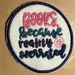 May include: A white round embroidered patch with a purple border. The patch says "Books, because reality is overrated" in pink, blue, and purple thread.