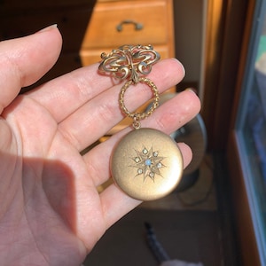 May include: A gold locket with a starburst design and a single pearl in the center. The locket is attached to a gold chain with a decorative butterfly charm.