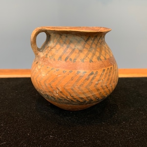 May include: A small, brown, ceramic jug with a handle. The jug has a geometric pattern painted in black on the surface.