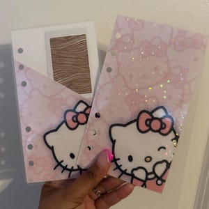 May include: Two translucent planner dashboard inserts featuring a repeating Hello Kitty pattern in soft pink. The sheets have a holographic glitter finish and pre-punched holes along the left side for binder insertion. One sheet includes a pocket.
