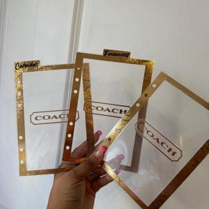 May include: Clear plastic planner dividers with gold foil trim and the word "COACH" printed in gold. The dividers are labeled "Calendar" and "Finances". The dividers have holes for binding.