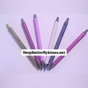 May include: A selection of glitter pens in shades of gold, purple, lavender, white, and blue, set against a pale purple backdrop. The pens feature silver details and the text "ShopButterflykisses.net".