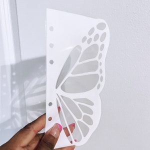May include: White plastic butterfly stencil with a detailed design. The stencil has a series of holes along the left edge.
