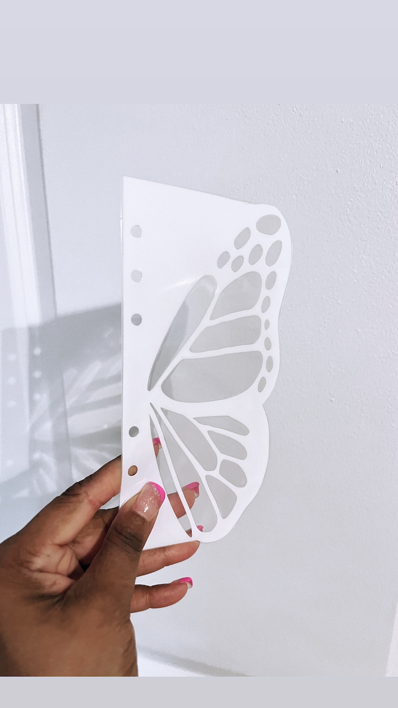 May include: White plastic butterfly stencil with a detailed design. The stencil has a series of holes along the left edge.