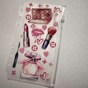May include: Clear cosmetic organizer with pink makeup-themed illustrations. Features a lipstick, eyeshadow palette, perfume bottle, makeup brush, mascara, hearts, and other decorative elements. The organizer has a zipper closure and is designed for storing beauty products.
