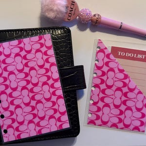 May include: A black leather planner with a pink and white patterned insert. A pink pen and a pink pom-pom pen topper are also visible. A clear plastic pocket with a pink and white patterned insert and a "TO DO LIST" card is also present.