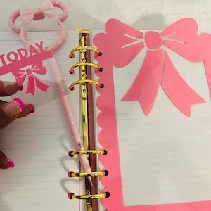 May include: Pink planner accessories including a pen, a decorative bow-shaped frame, and a clear overlay with the word "TODAY" and a bow graphic. The pen is pink with white polka dots. The planner has gold rings.