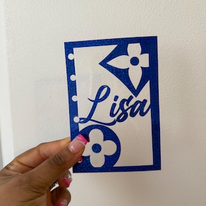 Peut inclure: Découpe en papier pailleté bleu avec un motif de fleur blanche et le nom "Lisa" écrit en cursive.