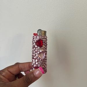 Pu&ograve; includere: Un accendino rosa con un corpo in plastica trasparente ricoperto di strass. L'accendino ha un design a forma di cuore rosso e una parte superiore argentata.