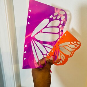 May include: Two butterfly stencils, one is a holographic pink and purple color and the other is orange. The stencils are held in a hand.