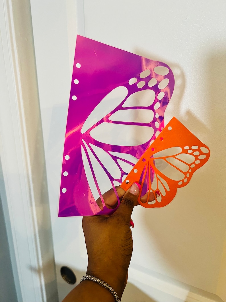 May include: Two butterfly stencils, one is a holographic pink and purple color and the other is orange. The stencils are held in a hand.