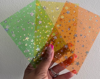May include: Four sheets of translucent paper with a holographic star pattern. The sheets are layered in shades of green, yellow, and orange.