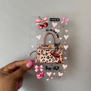 May include: A clear rectangular sheet featuring a graphic print of a leopard print handbag with a Coach logo, surrounded by pink hearts, red cherries with pink bows, and a black horse and carriage silhouette. The text Coach appears in multiple locations.
