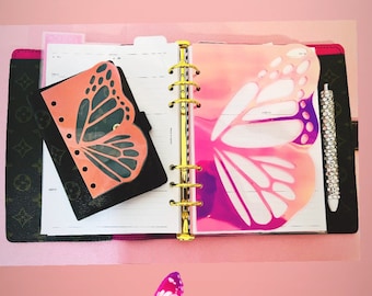 May include: A pink and white butterfly-shaped laminated dashboard insert for a planner or notebook. The insert is designed to be used as a decorative element and to help organize pages. The insert is shown in a planner with a gold ring binder.