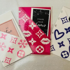 Puede incluir: Tres bolsillos de plástico transparente con diseños rosa, blanco y azul. Los bolsillos tienen un fondo blanco con flores rosa y blancas, labios rosas y un patrón Louis Vuitton rosa y blanco. El segundo bolsillo tiene un fondo blanco con corazones azules y un patrón Louis Vuitton azul y blanco. El tercer bolsillo tiene un fondo blanco con corazones azules y un patrón Louis Vuitton azul y blanco. Cada bolsillo tiene una tarjeta negra con texto blanco. El texto en las tarjetas dice "Be true", "Chase your dreams", y "Follow your heart".