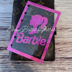 May include: Pink glitter sticker with a Barbie silhouette and the word "Barbie" in pink glitter letters. The sticker is on a brown leather planner.