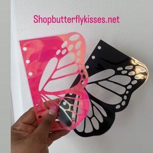 May include: Two butterfly-shaped stencils, one pink iridescent and one black, are shown on a white background. The stencils are designed for use with a planner or journal. The website Shopbutterflykisses.net is visible in the top center of the image.