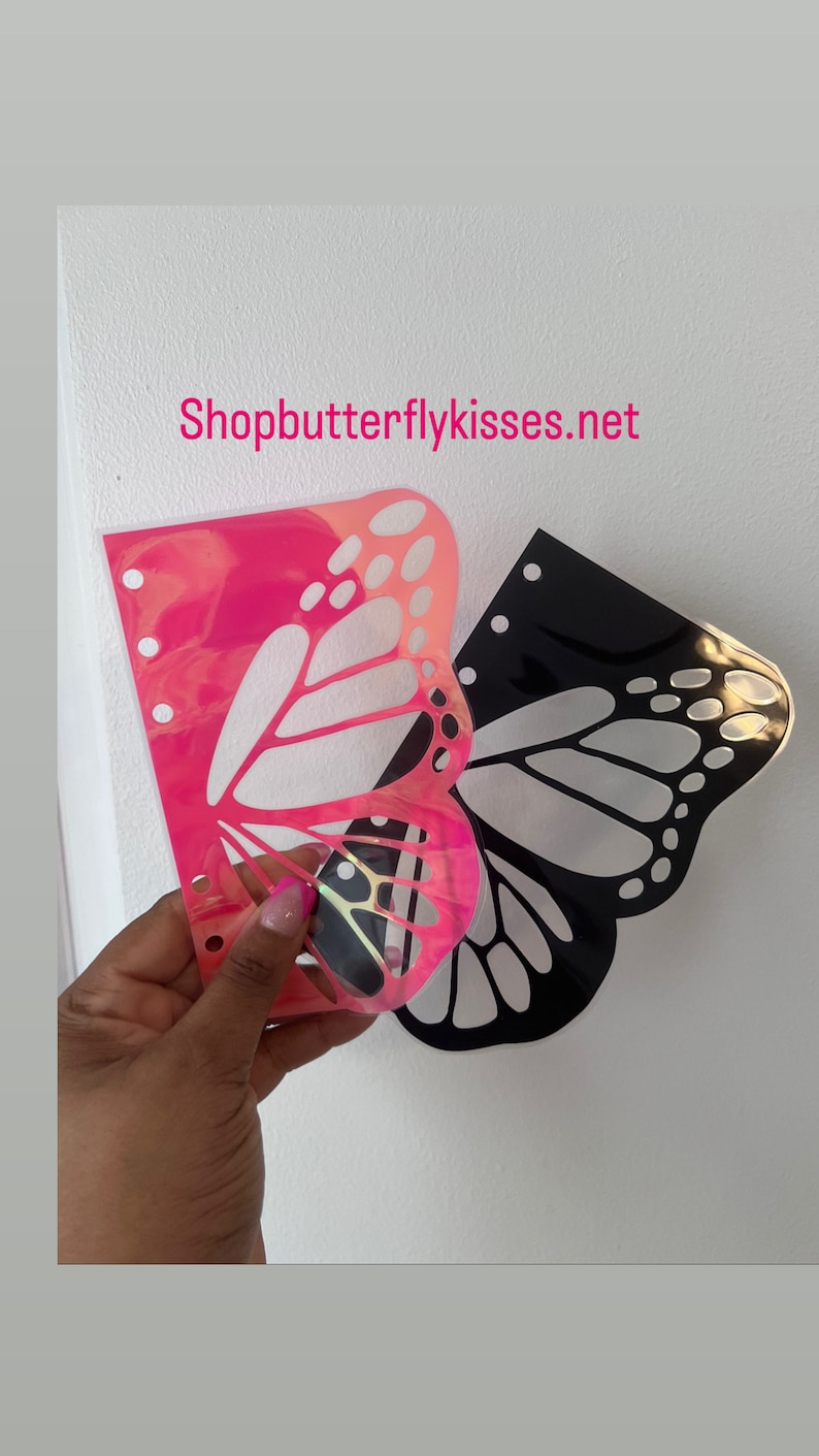 May include: Two butterfly-shaped stencils, one pink iridescent and one black, are shown on a white background. The stencils are designed for use with a planner or journal. The website Shopbutterflykisses.net is visible in the top center of the image.