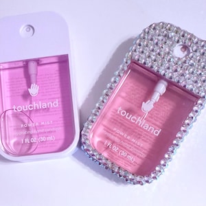 Może przedstawiać: Dwie różowe butelki z rozpylaczem do dezynfekcji rąk z białymi zakrętkami. Butelka po lewej stronie jest gładka, a butelka po prawej stronie jest pokryta tęczowymi kryształkami. Na obu butelkach widnieje napis "touchland POWER MIST Hydrating Hand Sanitizer 1 FL OZ (30 mL)" nadrukowany z przodu.
