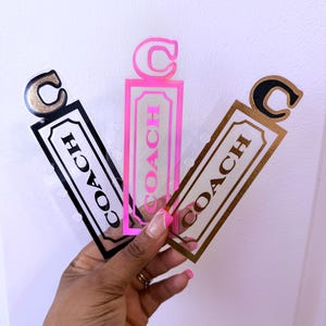 Op de afbeelding: Drie Coach-merk bladwijzers in zwart, roze en goud. Elke bladwijzer heeft een rechthoekig label met het woord "COACH" erop gedrukt. De bovenkant van elke bladwijzer heeft een "C"-vorm.
