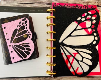 May include: A brown and pink butterfly-themed planner and notebook set. The planner features a brown cover with a pink butterfly cutout. The notebook has a black cover with a white and red butterfly wing design. Gold rings bind the pages.
