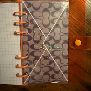 May include: An open orange planner with a grid-lined page and a patterned insert. The insert features a repeating logo design in shades of gray and white, with a holographic overlay. The planner has orange metal rings and a snap closure.