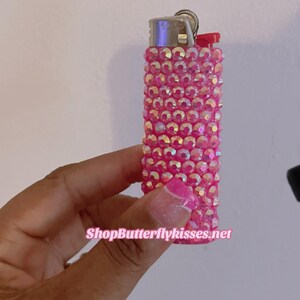 Pu&ograve; includere: Accendino rosa ricoperto di strass iridescenti. L'accendino ha una parte superiore argentata e una finestra in plastica trasparente. Il testo "ShopButterflykisses.net" &egrave; visibile nella parte inferiore dell'immagine.