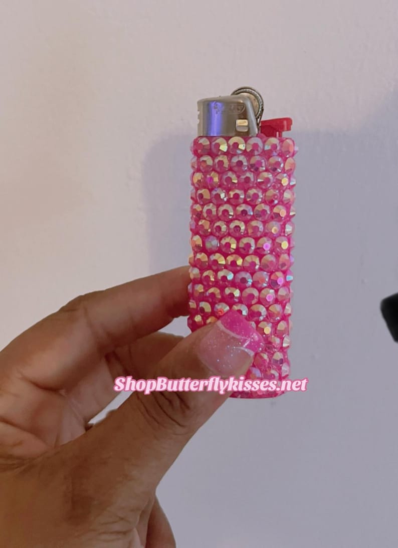 Pu&ograve; includere: Accendino rosa ricoperto di strass iridescenti. L'accendino ha una parte superiore argentata e una finestra in plastica trasparente. Il testo "ShopButterflykisses.net" &egrave; visibile nella parte inferiore dell'immagine.