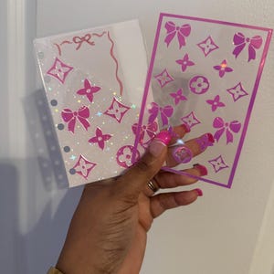 Puede incluir: Dos insertos de planificador transparentes con patrones de lazo y logotipo rosas. Uno es un bolsillo con un diseño de lazo en la parte superior. El otro es una hoja transparente con un borde rosa y varios diseños de logotipo y lazo rosas.