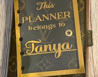 May include: A personalized planner with a brown and gold design. The planner cover features a gold-framed insert with the text "This PLANNER belongs to Tanya" in gold lettering. The planner has a dark brown background with a repeating pattern.