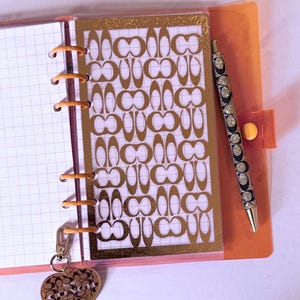 Peut inclure: Un agenda orange avec un motif doré du logo Coach sur un transparent. Un stylo assorti avec un motif noir et argenté est posé à côté de l'agenda. Un charm en forme de cœur avec le logo Coach est suspendu aux anneaux.