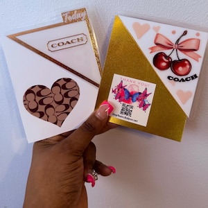 Peut inclure: Deux cartes de la marque Coach sont tenues. L'une présente un motif en forme de cœur, l'autre un motif de cerises. Les deux ont des accents dorés et le logo Coach. Un autocollant avec des papillons et le texte "Thank You!" est visible.