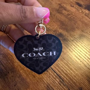May include: A heart-shaped black keyring featuring the Coach New York logo and horse and carriage emblem in white, with a repeating dark grey signature pattern and a gold-toned metal lobster clasp attachment.