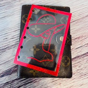 May include: A brown and tan planner with a red-outlined illustration of a woman wearing a hat. The illustration is on a clear plastic sheet with a red glitter border. The planner is closed and has a snap closure.