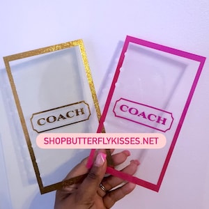 Może przedstawiać: Dwa przezroczyste etui na karty marki Coach. Jeden ma złotą obwódkę i złote logo Coach. Drugi ma różową obwódkę i różowe logo Coach. Na różowym banerze wydrukowano stronę internetową shopbutterflykisses.net.