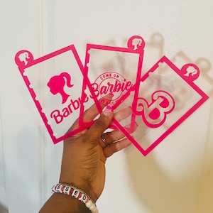 May include: Four pink plastic stencils with the word "Barbie" and a silhouette of a Barbie doll. The stencils are held in a hand wearing a beaded bracelet.
