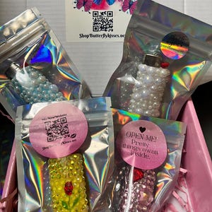 Pu&ograve; includere: Quattro piccoli accendini decorati in sacchetti di plastica trasparente individuali. Gli accendini sono decorati con strass e perle. I sacchetti sono su una superficie rosa. Il testo "OPEN ME! Pretty things await inside." &egrave; su un adesivo rosa a forma di cuore.