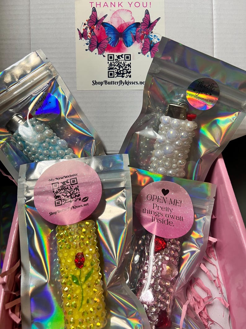 Pu&ograve; includere: Quattro piccoli accendini decorati in sacchetti di plastica trasparente individuali. Gli accendini sono decorati con strass e perle. I sacchetti sono su una superficie rosa. Il testo "OPEN ME! Pretty things await inside." &egrave; su un adesivo rosa a forma di cuore.