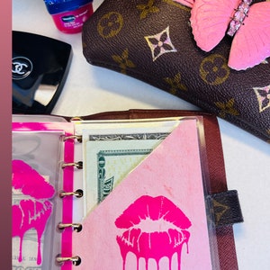 May include: A pink and brown leather planner with a clear plastic pocket. The pocket contains a pink piece of paper with a pink dripping lipstick design and the text "Shop Butterflykisses".