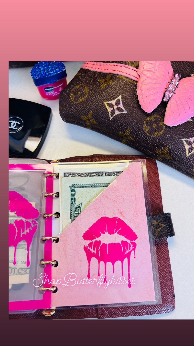 May include: A pink and brown leather planner with a clear plastic pocket. The pocket contains a pink piece of paper with a pink dripping lipstick design and the text "Shop Butterflykisses".
