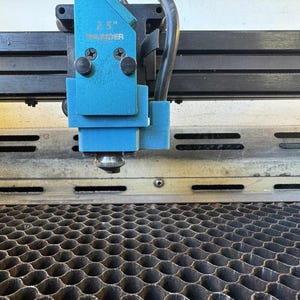 May include: Close-up of a laser cutter machine. A bright blue laser head is mounted on a black rail. The head has the text "2.5" and "SHENDER" printed on it. Below the head is a metal surface with horizontal slots, and a honeycomb grid.
